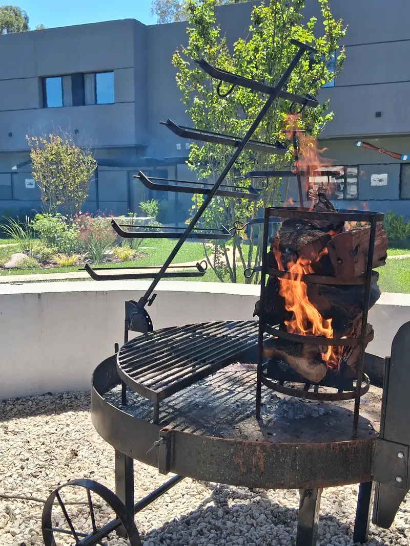 Fogón con parrilla y asador de hierro encendido, rodeado de piedras, con el jardín y los dúplex de fondo.