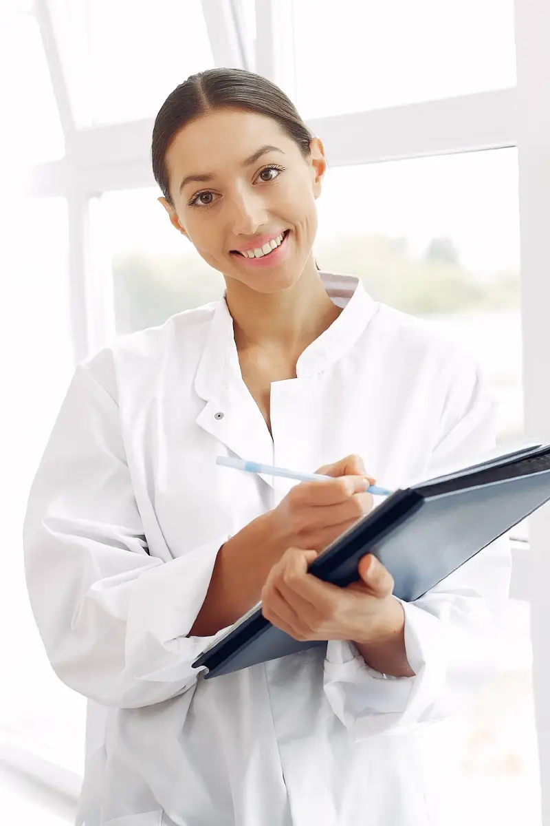 Profesional de la salud con guardapolvo blanco, sonriendo mientras toma notas en una carpeta.